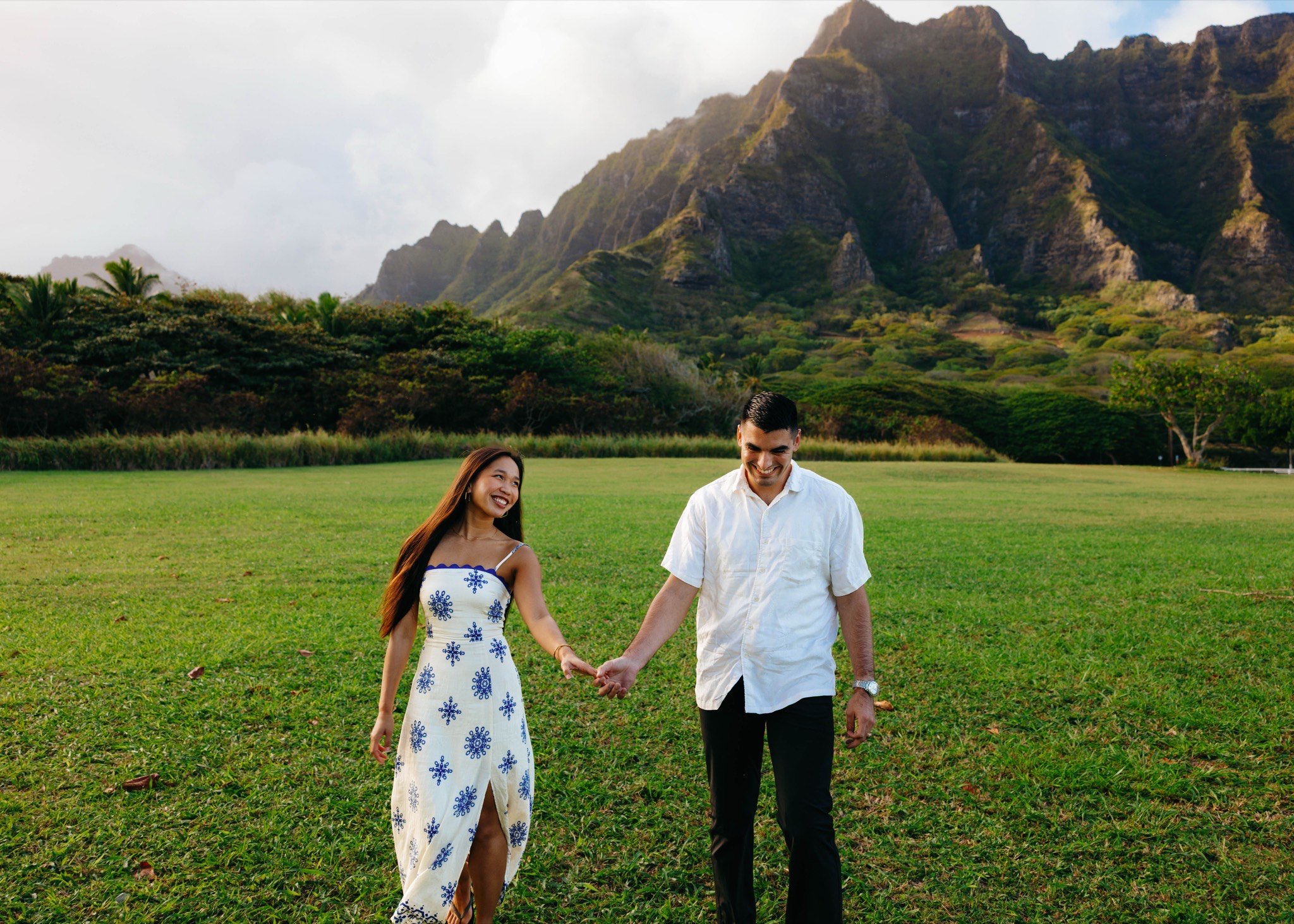 Abigail and Edmund walking hand in hand across the lawn at Kualoa, the Koʻolau ridge rising behind them.